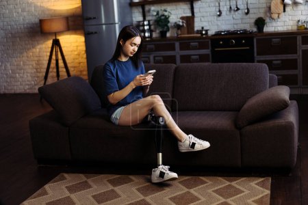 Photo for Woman with disability sits on sofa and types messages on smartphone. Attractive lady with prosthetic leg engaged in lively chat with boyfriend on mobile phone - Royalty Free Image