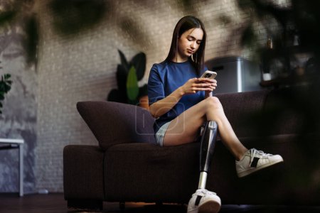 Photo for Woman with disability sits on sofa and types messages on smartphone. Attractive lady with prosthetic leg engaged in lively chat with boyfriend on mobile phone - Royalty Free Image