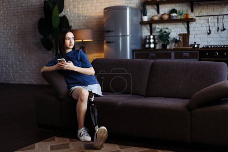 Photo for Young woman with disability shares stories with friend using phone. Lady with prosthetic leg in casual attire sits on soft couch and types messages on smartphone - Royalty Free Image