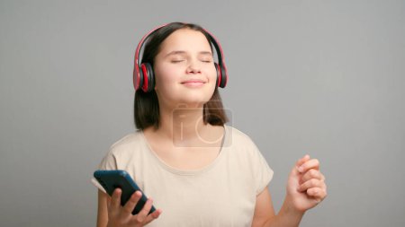 Téléchargez les photos : Femme adolescente latine souriante écoutant le podcast e-book musique chanson chanteur groupe rock dans les écouteurs casque, en choisissant la bande sonore sur téléphone portable isolé sur fond gris. - en image libre de droit