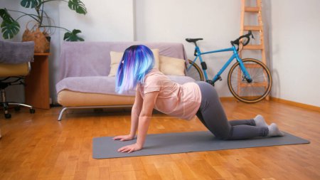 Young plus size latin hispanic teenager girl doing stretching when vacuum cleaner robot working around. Cozy home interior with indoor plants. Online fitness, urban jungle concept.