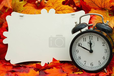 Foto de Signo en blanco con un reloj despertador y hojas de otoño para su cambio de hora o mensaje de hora de otoño - Imagen libre de derechos
