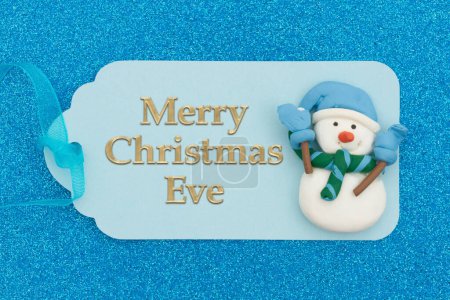 Photo for Merry Christmas Eve greeting on teal holiday gift tag with a snowman on glitter paper with a ribbon - Royalty Free Image