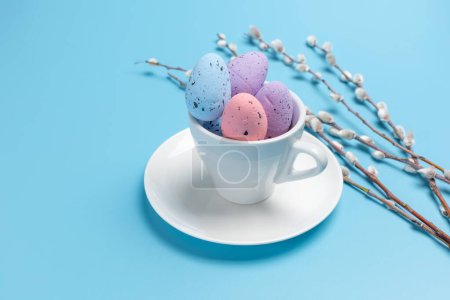 Porcelain cup with colored Easter eggs on a saucer and willow branches with catkins on the blue background. Top view.
