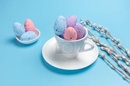 Foto de Copa de porcelana con huevos de Pascua de colores en un platillo y ramas de sauce con amentos en el fondo azul. Vista superior. - Imagen libre de derechos