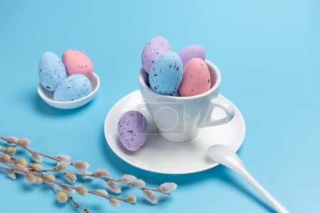 Foto de Copa de porcelana con huevos de Pascua de colores en un platillo, una cuchara y ramas de sauce con amentos en el fondo azul. Vista superior. - Imagen libre de derechos
