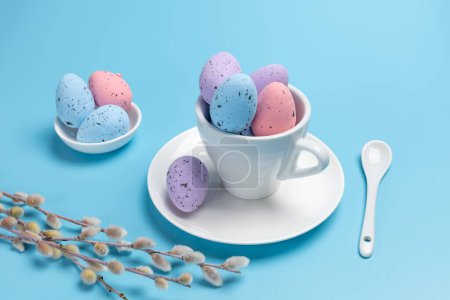 Foto de Copa de porcelana con huevos de Pascua de colores en un platillo, una cuchara y ramas de sauce con amentos en el fondo azul. Vista superior. - Imagen libre de derechos