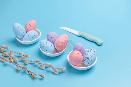 Foto de Platillos de porcelana con huevos de Pascua de colores, un cuchillo y ramas de sauce con amentos en el fondo azul. Vista superior. - Imagen libre de derechos