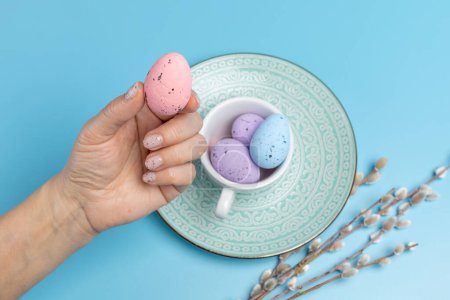 Female hand and a porcelain cup with colored Easter eggs. Willow branches with catkins on the blue background. Top view.