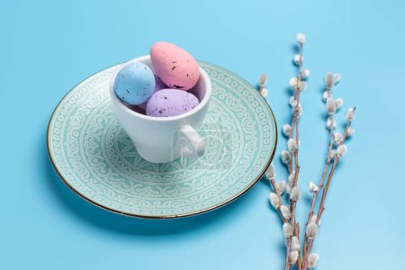 Copa de porcelana con huevos de Pascua de colores en un plato y ramas de sauce con amentos en el fondo azul. Vista superior.