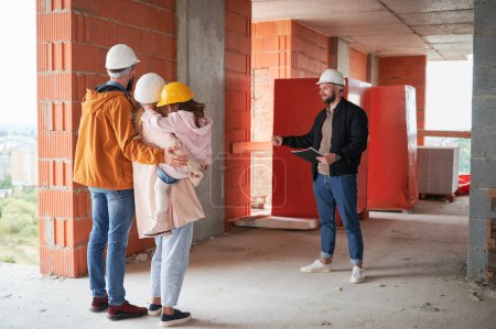 Man and woman with child standing inside apartment building under construction and talking with builder. Family future homeowners discussing building process with construction worker.