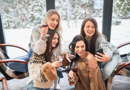 Young women enjoying winter weekends inside contemporary barn house. Four girls having fun, taking selfies on phone and drinking wine.
