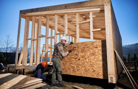 Foto de Carpenters hammering nail into OSB panel on the wall of future cottage. Men workers building wooden frame house. Carpentry and construction concept. - Imagen libre de derechos