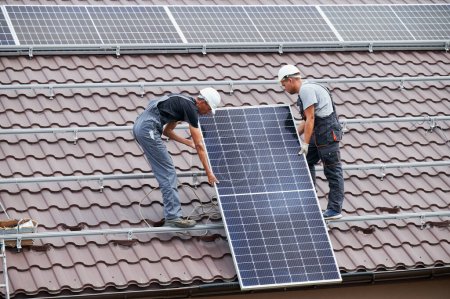 Téléchargez les photos : Des techniciens hommes soulevant des modules solaires photovoltaïques sur le toit de la maison. Ingénieurs dans les casques de construction système de panneaux solaires à l'extérieur. Concept d'énergies alternatives et renouvelables. - en image libre de droit