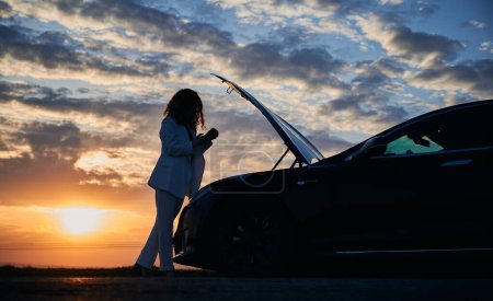 Silhouette der Frau Suchdienst der Pannenhilfe für die Behebung von Problemen mit Elektroauto unter der Motorhaube mit Smartphone-Technologie während der Sonnenuntergangsfahrt, Fahrerin haben Online-Unterstützung