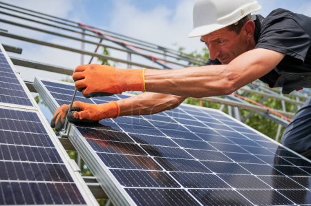 Close up of man installer placing solar module on metal rails. Male worker mounting photovoltaic solar panel system outdoors, wearing construction helmet and work gloves.