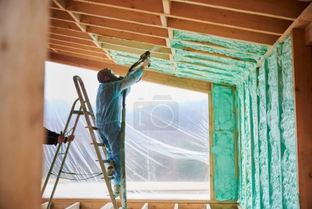 Männliche Bauarbeiter isolieren Holzrahmenhaus. Arbeiter versprüht Polyurethanschaum im Inneren der zukünftigen Hütte, steht auf einer Leiter und benutzt eine Mehrkomponentenpistole. Bau- und Dämmkonzept.