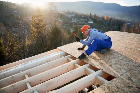 Carpenter hammering nail into OSB panel on the roof top of future cottage at sunrise. Man worker building wooden frame house. Carpentry and construction concept.