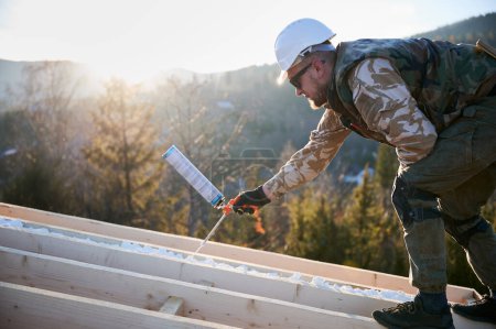 Photo for Male builder doing thermal insulation on roof of wooden frame house in the evening. Man worker spraying polyurethane foam on rooftop of future cottage. Construction and insulation concept. - Royalty Free Image