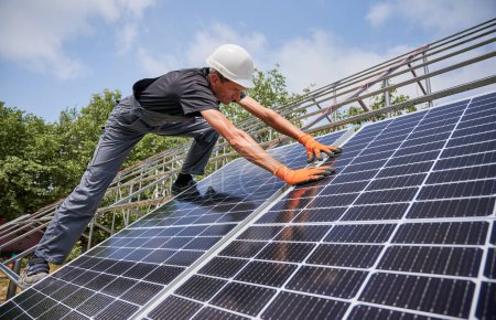 Homme en casque de sécurité et salopettes de travail bâtiment photovoltaïque système de panneaux solaires à l'extérieur. Travailleur masculin en gants orange plaçant des modules solaires sur des rails métalliques. Concept de sources d'énergie alternatives.