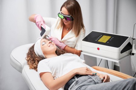 Foto de Smiling woman in protective goggles receiving laser facial treatment in beauty salon. Female cosmetologist using diode laser scanner device while performing resurfacing skincare procedure. - Imagen libre de derechos