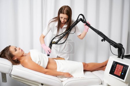 Photo for Doctor cosmetologist using erbium ablative laser machine while performing lifting procedure on female abdomen. Young woman receiving laser treatment in cosmetology clinic or beauty salon. - Royalty Free Image