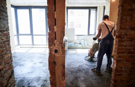 Back view of male worker using troweling machine while screeding floor in apartment under renovation. Man finishing concrete surface with floor screed grinder machine in room with large windows.