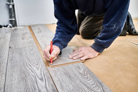 Photo for Close up of man construction worker with pencil in hand drawing mark on laminate wooden plank. Male worker preparing laminate boards for floor installation in apartment under renovation. - Royalty Free Image