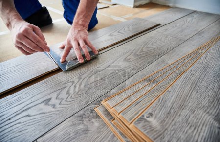 Man preparing laminate plank for floor installation in apartment under renovation. Close up of male worker using metal construction ruler and pen while drawing line on laminate flooring board.
