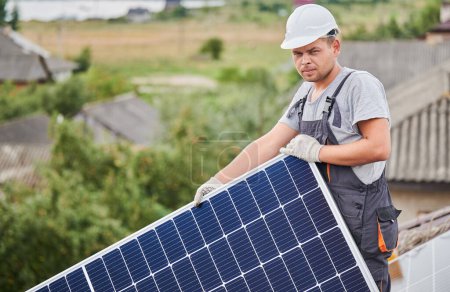 Portrait of man worker mounting photovoltaic solar moduls on roof of house. Electrician in helmet installing solar panel system outdoors. Concept of alternative and renewable energy.