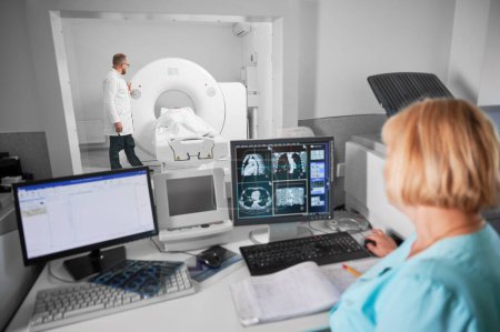Medical computed tomography or MRI scanner. Back view of blonde nurse sitting at computer, doctor standing, making MRI. Specialists working in clinic. Concept of modern diagnostics.