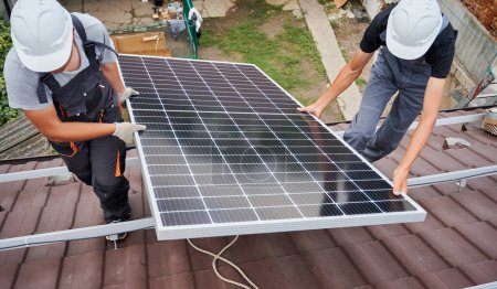 Téléchargez les photos : Les hommes montent soulever des modules solaires photovoltaïques sur le toit de la maison. Electriciens dans les casques montage système de panneaux solaires à l'extérieur. Concept d'énergies alternatives et renouvelables. - en image libre de droit