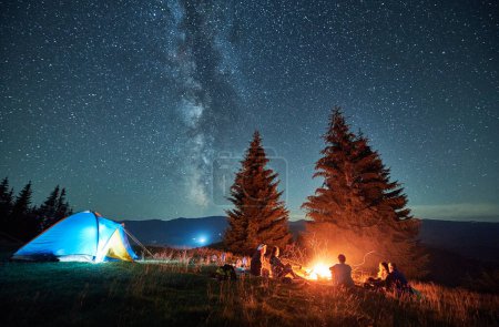 Nächtliches Zelten in den Bergen unter Sternenhimmel. Eine Gruppe Touristen, die sich in der Nähe des Zeltplatzes ausruhen, ein brennendes Lagerfeuer und ein beleuchtetes Zelt. Tourismus-, Wander- und Erlebniskonzept.