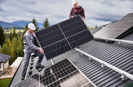 Foto de Ingenieros construyendo sistema de paneles solares en el techo de la casa. Hombres trabajadores en cascos que llevan módulo solar fotovoltaico al aire libre. Concepto de energía alternativa y renovable. - Imagen libre de derechos