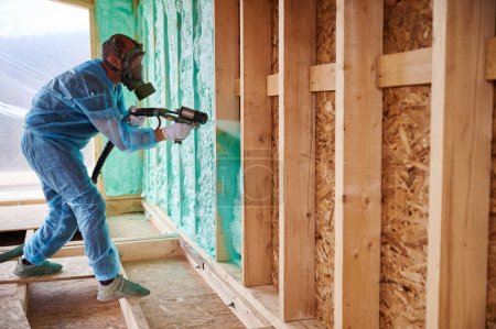 Männliche Bauarbeiter isolieren Holzrahmenhaus. Mann Arbeiter sprüht Polyurethan-Schaum innerhalb der zukünftigen Hütte, mit Mehrkomponenten-Pistole. Bau- und Dämmkonzept.