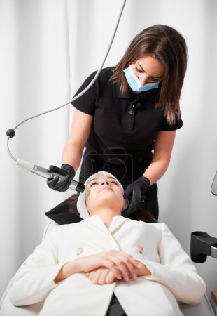 Photo for Female cosmetologist in medical mask using professional beauty device while performing face lifting procedure in cosmetology clinic. Woman receiving radiofrequency facial treatment in clinic. - Royalty Free Image