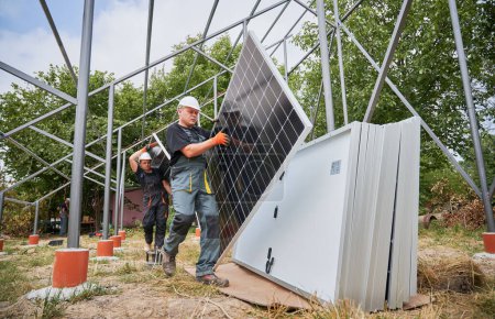 Workers carrying solar panels for installing in field. European men wearing workwear and helmets. Photo-voltaic collection of modules as a PV panel. Array as a system of photo-voltaic panels.