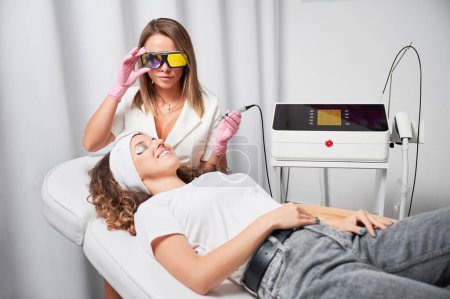 Photo for Female beautician in protective glasses using diode laser device while performing skin rejuvenation procedure in beauty salon. Young woman in silver goggles lying on daybed and smiling. - Royalty Free Image
