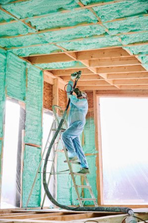 Photo for Male builder insulating wooden frame house. Man worker spraying polyurethane foam inside of future cottage, using plural component gun. Construction and insulation concept. - Royalty Free Image
