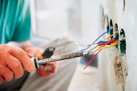 Photo for Close up of man in work gloves using electric repair solder welding tool while installing electrical cables in apartment under renovation. Male electrician working with electric heater soldering iron. - Royalty Free Image
