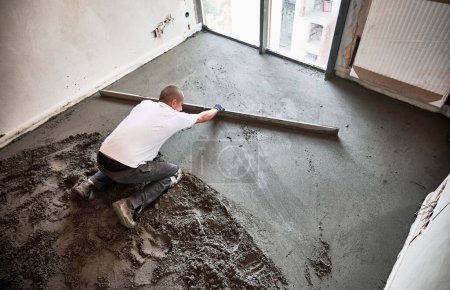 Draufsicht eines männlichen Bauarbeiters, der eine Estrichschiene auf den mit Sand-Zement-Gemisch bedeckten Boden legt. Mann glättet und nivelliert Fläche mit gerader Kante beim Estrichboden in Wohnung.