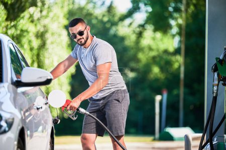 Photo for Driver with gasoline pump refilling car gas tank. Confident man refueling his luxury white auto. Man in casual clothes and sunglasses at modern gas station. - Royalty Free Image