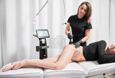 Photo for Doctor cosmetologist using radiofrequency microneedling device while performing skin tightening treatment on female hip. Smiling woman lying on medical chair during stretch marks removal procedure. - Royalty Free Image