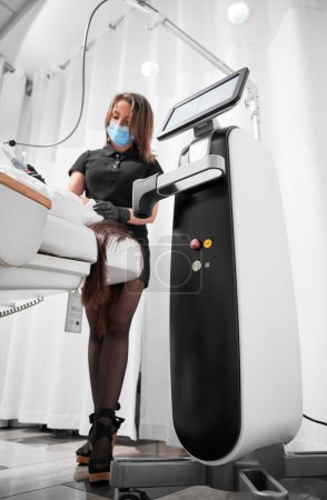 Photo for Full length of female cosmetologist in medical mask using professional equipment radiofrequency machine in cosmetology clinic. Doctor aesthetician performing radiofrequency facial treatment for woman. - Royalty Free Image