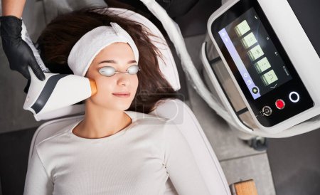 Photo for Top view of doctor cosmetologist using diode laser scanner device while performing resurfacing skincare procedure. Woman in protective glasses getting rejuvenation facial treatment. - Royalty Free Image