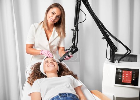 Photo for Female doctor cosmetologist using erbium ablative laser machine while performing resurfacing procedure. Smiling woman lying on daybed and receiving facial skincare treatment in beauty salon. - Royalty Free Image