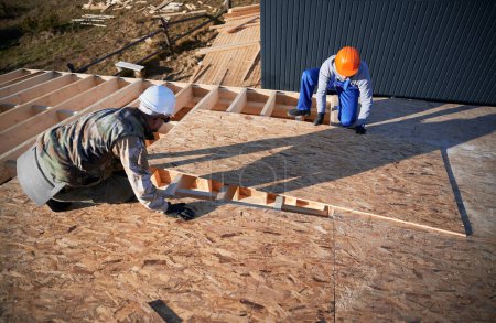 Téléchargez les photos : Carpenters mounting wooden OSB board on rooftop of future cottage. Men workers building wooden frame house. Carpentry and construction concept. - en image libre de droit