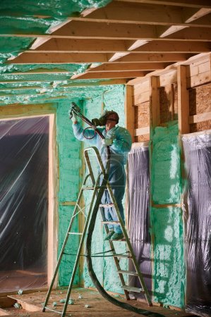 Male builder insulating wooden frame house. Man worker spraying polyurethane foam inside of future cottage, standing on ladder, using plural component gun. Construction and insulation concept.