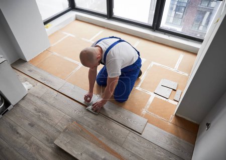 Top view of man in workwear using metal construction ruler and pen while drawing line on laminate flooring board. Male worker preparing laminate plank for floor installation in flat under renovation.