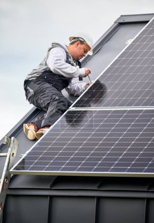 Man technician mounting photovoltaic solar panels on roof of house. Engineer in helmet installing solar module system with help of hex key. Concept of alternative, renewable energy.